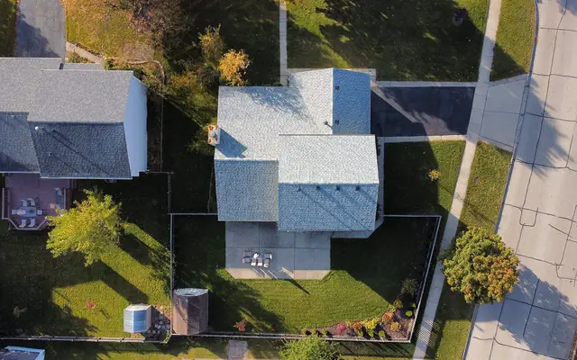 Top-down view of a house with a light grey shingle roof, a patio and a fenced back lawn with two small sheds